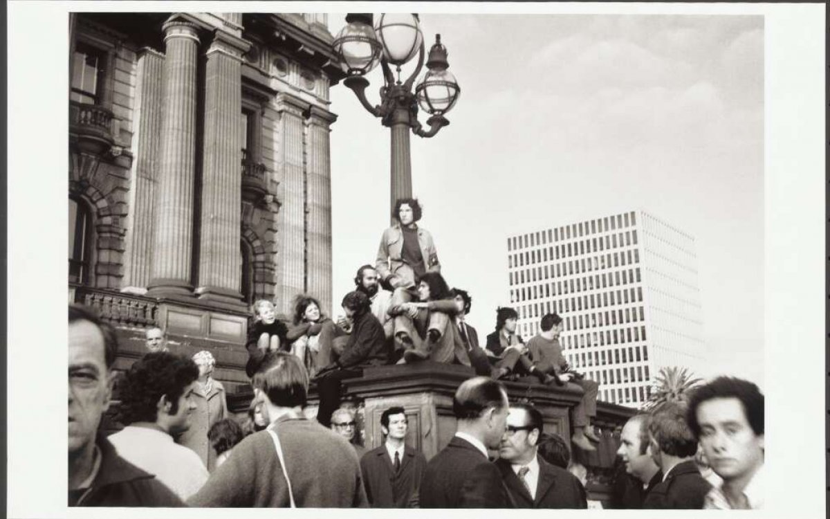 A black and white photo of tens of people gathered on top of and around a large light  on a large stone plinth in front of Victoria's Parliament House.