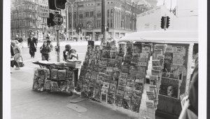 A black and white image of a newspaper seller sitting behind a bench table. There are several stands of magazines and newspapers with many different issues and titles. The stand is on the corner of a busy street in Sydney. In the backgroun the Queen Victoria Building can be seen. People are crossing the road.