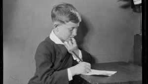 A black and white portrait of a young boy sitting at a desk. He is writing on a blank piece of paper.