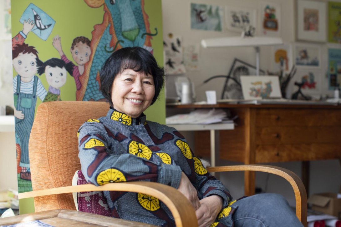 A person sitting in a chair with their arm resting on the chair's arm. Behind them is a banner with the image of a dragon and three children on it. In the background is a desk and wall covered with illustrations and artworks.