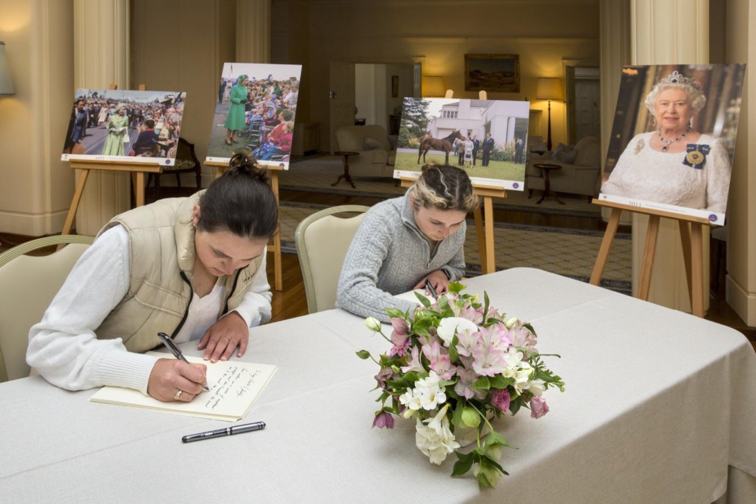 Two women sitting at a table writing on pieces of paper. There are images of Queen Elizabeth II on easels in the background.