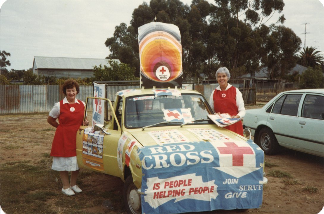 Two ladies in nurses uniforms standing either side of an old car that is covered in Red Cross logos and images.