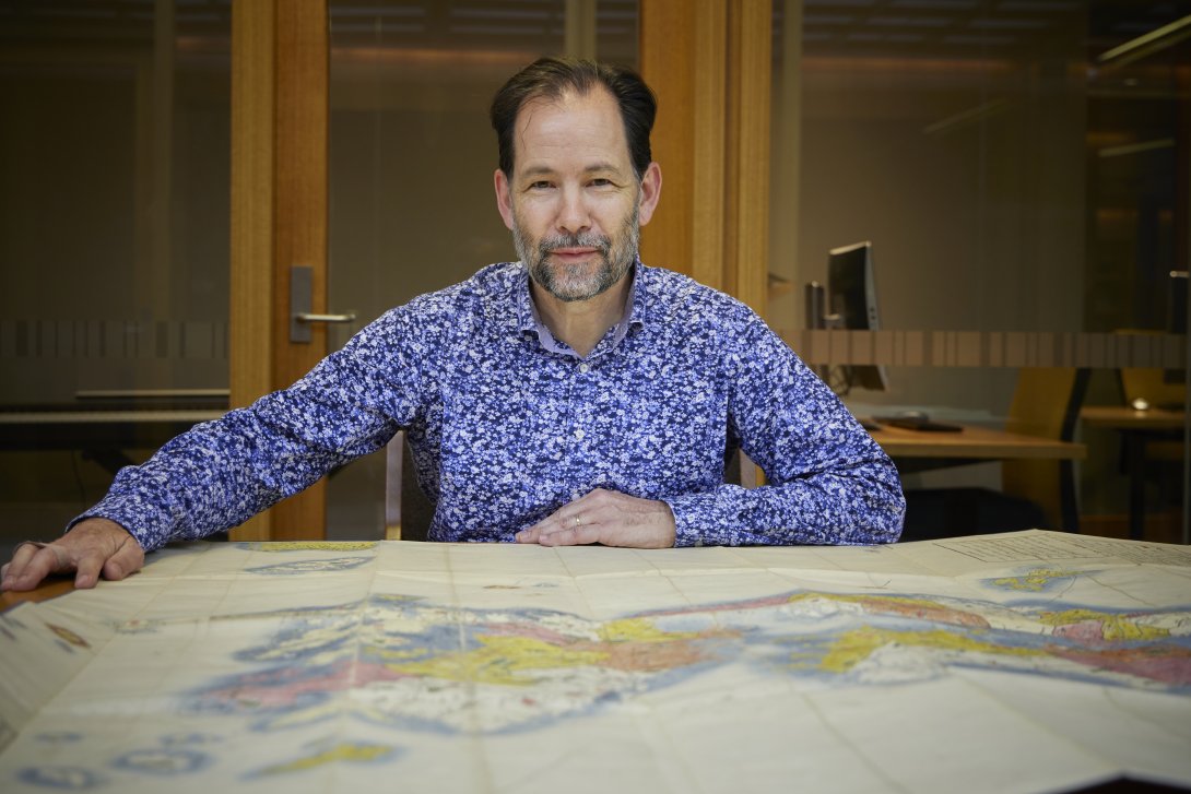 Man with brown hair and a grey and brown beard sitting at a table with a large map spread over it